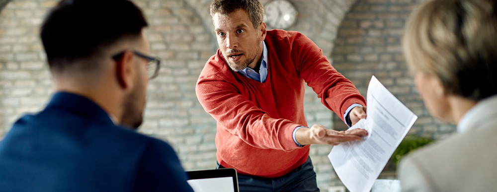 Ein Mann in einem roten Pullover hält energisch ein Dokument hoch und spricht gestikulierend mit zwei Geschäftspartnern in einem Meetingraum. Szene eines geschäftlichen Konflikts.