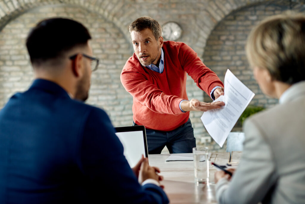 Ein Mann in einem roten Pullover hält energisch ein Dokument hoch und spricht gestikulierend mit zwei Geschäftspartnern in einem Meetingraum. Szene eines geschäftlichen Konflikts.